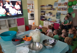 Dzieci siedzą i oglądają na ekranie interaktywnym film edukacyjny o kiszeniu kapusty.