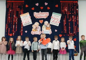 Dzieci stoją na scenie, chłopiec mówi wiersz i trzyma duże czerwone serce. W tle dekoracja z okazji Dnia Babci i Dziadka