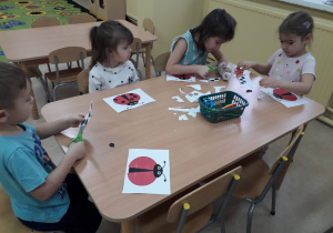 Dzieci przy stoliku ozdabiają papierowe biedronki kropkami.