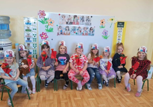 Dziewczynki z grupy młodszej siedzą na krzesełkach i pozują do zdęcia. W tle tematyczny napis "Dzień Kobiet".