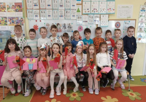 Grupa starsza pozuje do zdjęcia. Dziewczynki trzymają kwiatki i laurki.