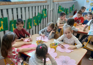 Dzieci siedzą przy stolikach i malują kwiaty z papieru