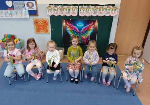 Dziewczynki z gr 3-4 latków siedzą z kwiatami na tle dekoracji