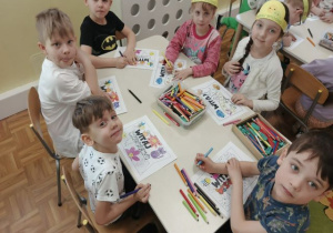 Dzieci siedzą przy stoliku i kolorują chorągiewki na powitanie wiosny.