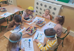Dzieci siedzą przy stoliku i kolorują chorągiewki na powitanie wiosny.