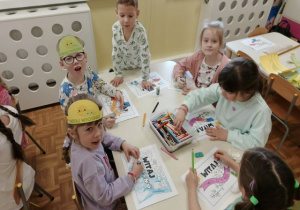 Dzieci siedzą przy stoliku i kolorują chorągiewki na powitanie wiosny.