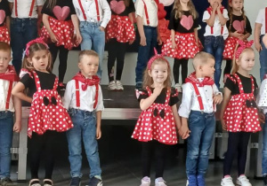 Dziewczynki ubrane w czerwone spódniczki w białe grochy i czarne bluzki, chłopcy w białych koszulach, spodniach i czerwonych szelkach śpiewają piosenkę dla rodziców.