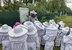 Dzieci ubrane w kombinezony pszczelarskie słuchają pana pszczelarza opowiadającego o cyklu rozowju pszczół.