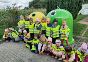Przedszkolaki pozują do zdjęcia, w tle widać pojeminki do segregacji odpadów.