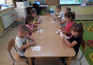 Dzieci przy stoliku wycinają cyfry i układają z nich numery alarmowe.