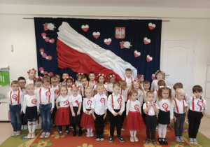 Przedszkolaki śpiewają hymn na tle biało-czerwonej flagi.