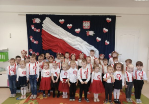 Przedszkolaki śpiewają hymn na tle biało-czerwonej flagi.