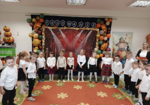 Grupa starsza stoi w półkolu na tle dekoracji i śpiewa piosenkę.