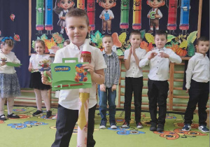 Chłopiec pozuje do zdjęcia z ołówkiem i dyplomem. W tle dzieci biją brawo.