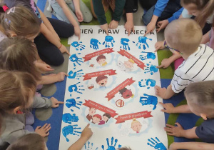 Dzieci siedzą na dywanie. W środku leży plakat.