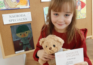 Dziewczynka pozuje do zdjęcia z misiem i dyplomem. W tle praca konkursowa.