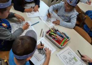 Przedszkolaki siedzą przy stoliku i kolorują obrazki.
