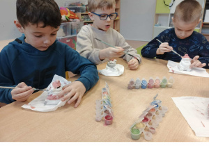 Dzieci siedzą przy stoliku i malują ceramiczne skarbonki Mikołaje