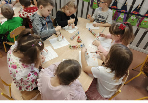 Dzieci siedzą przy stoliku i malują ceramiczne skarbonki Mikołaje