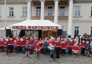Przedszkolaki ubrane w czerwone peleryny i czapki śpiewają świąteczne piosenki.
