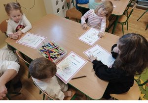 Kilkoro dzieci z grupy 3, 4 latków siedzi przy stoliku w sali przedszkolnej, rysują kredkami na kartce wymarzone prezenty.