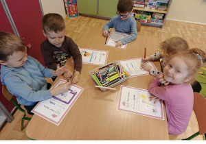 Następna grupa 3, 4 latków siedzi przy stoliku w sali przedszkolnej, rysują kredkami na kartce wymarzone prezenty.