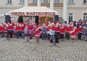 Dzieci ubrane w czerwone peleryny i czapki świąteczne śpiewają piosenki.