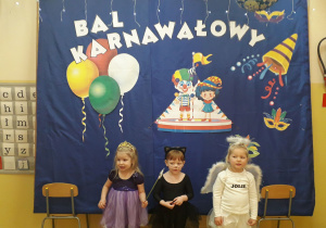 Dziewczynki w strojach karnawałowych pozują do zdjęcia. W tle karnawałowa dekoracja.