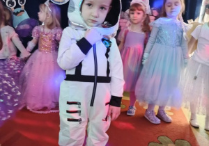 Chłopiec w stroju astronauty, w oddali tańczące dziewczynki przebrane za księżniczki.
