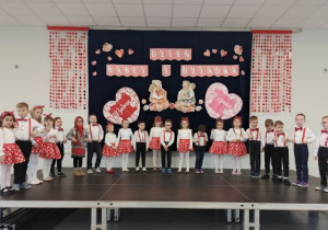 Dzieci ze starszej grupy ustwione w półkolu stoją na scenie i śpiewają piosenkę.