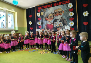 Dzieci śpiewają piosenkę trzymając serca na patyczkach. W tle dekoracja.