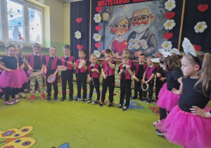 Dziewczynki stoją po bokach i śpiewają piosenkę. Chłopcy w środku grają na instrumentach.