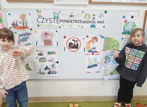 Dwie dziewczynki stoją przy tablicy z ilustracjami przedstawiającymi zanieczyszczenia powietrza i sposoby jak je ograniczać