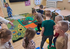 Przedszkolaki spacerują po ścieżce sensorycznej wykonanej z kamieni sensorycznych, kasztanów, klocków i folii bąbelkowej.