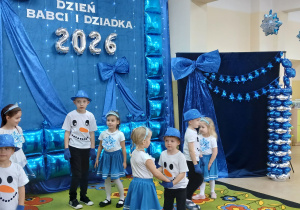 Dzieci w biało niebieskich strojach na tle dekoracji prezentują taniec.