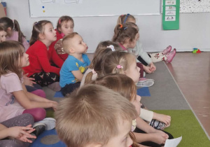 Dzieci siedzą na dywanie i oglądają prezentację, w tle tablica z informacjami na temat bezpieczeństwa w sieci