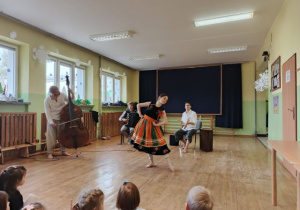 Panowie grają na instumentach a pani ubrana w łowicki strój prezentuje taniec.
