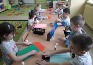 Dzieci przy stolikach projektują i sklejają domki z figur geometycznych.