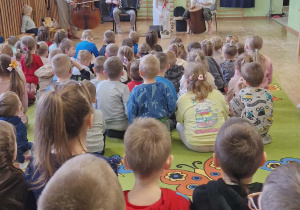 Dzieci siedzą na dywanie. Przed nimi muzycy grają na instrumentach i pani ubrana w białą sukienkę trzyma koszyk z ziołami