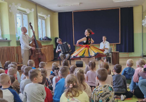Dzieci siedzą na dywanie. Przed nimi muzycy grają na instrumentach i pani ubrana w łowicki strój tańczy