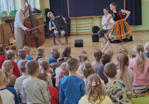 Dzieci siedzą na dywanie. Przed nimi muzycy grają na instrumentach i pani ubrana w łowicki strój tańczy
