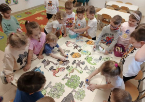 Przedszkolaki stoją dookoła stołu i przyklejają obrazki dinozaurów na dużym brystolu.
