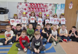 Dzieci pozują do zdjęcia grupowego na tle dekoracji. Dziewczynki trzymają laurki od chłopców