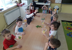 Dzieci przy stolikach prezentują wykonany napój z wody, owoców i świeżych ziół.