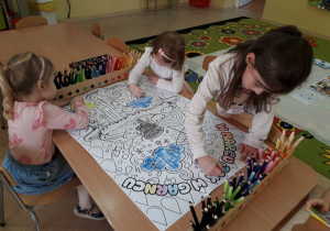 Dziewczynki przy stoliku kolorują duży plakat "W marcu jak w garncu".