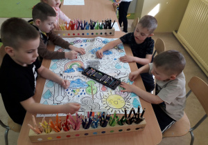 Chłopcy przy stoliku kończą kolorować duży plakat "W marcu jak w garncu".