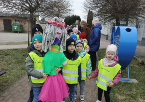 Przedszkolaki ubrane w kamizelki odblaskowe, ustawione parami przygotowują się do spaceru. Chłopiec trzyma marzannę.