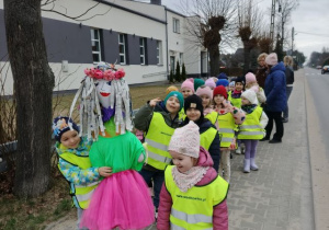 Dzieci ubrane w kamizelki odblaskowe spacerują po chodniku, chłopiec trzyma marzannę.