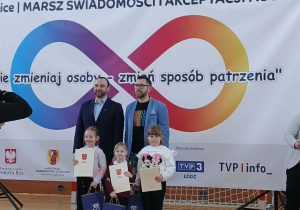 Dzieci przedszkolne z wręczonymi dyplomami i nagrodami stoją z przedstawicielami wydarzenia na tle dekoracji.