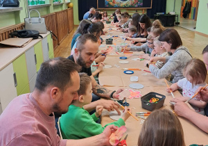 Dzieci z rodzicami siedzą przy stołach i wykonują podstawki z materiałów ekologicznych.